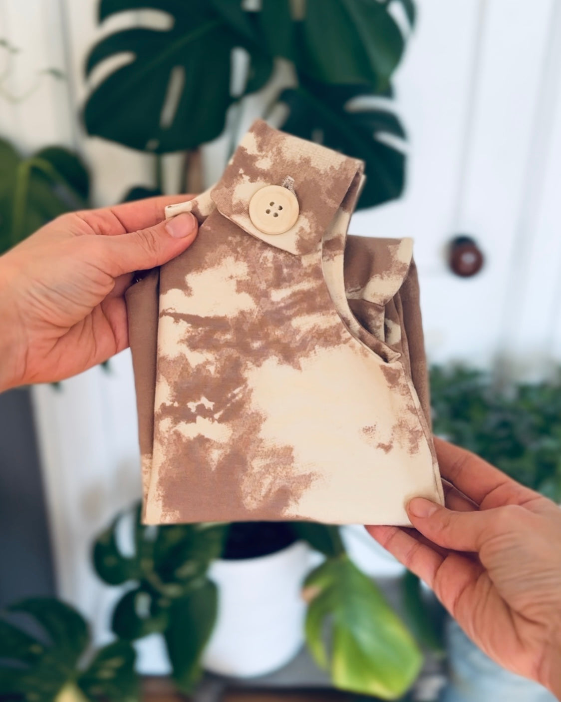 Mauve tie-dye romper folded being held by hands in front of a blurred plant backdrop