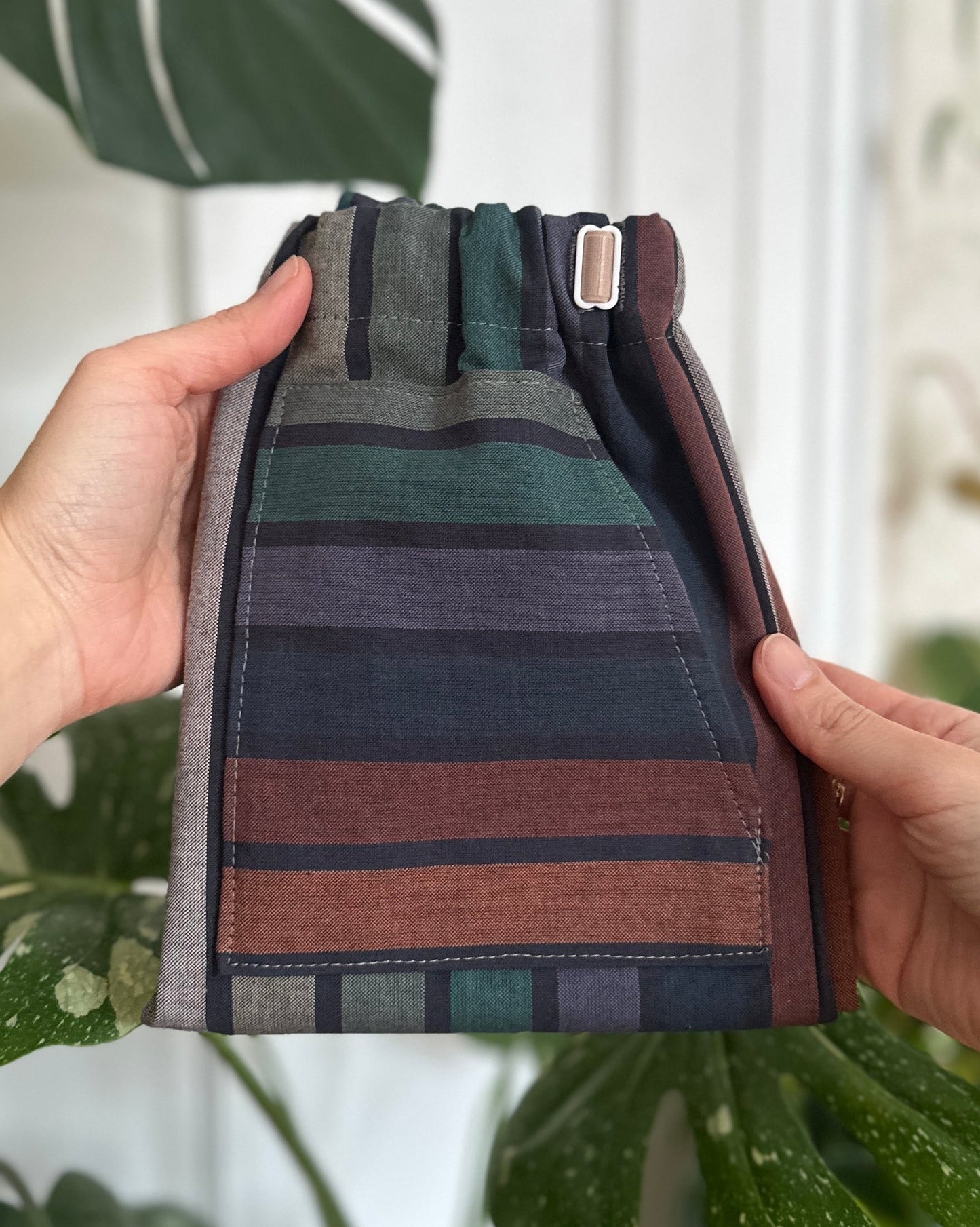 Folded striped fabric with green, brown, and gray colors held by a person against a blurred indoor plant background.