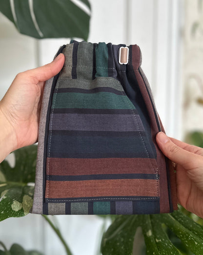 Folded striped fabric with green, brown, and gray colors held by a person against a blurred indoor plant background.