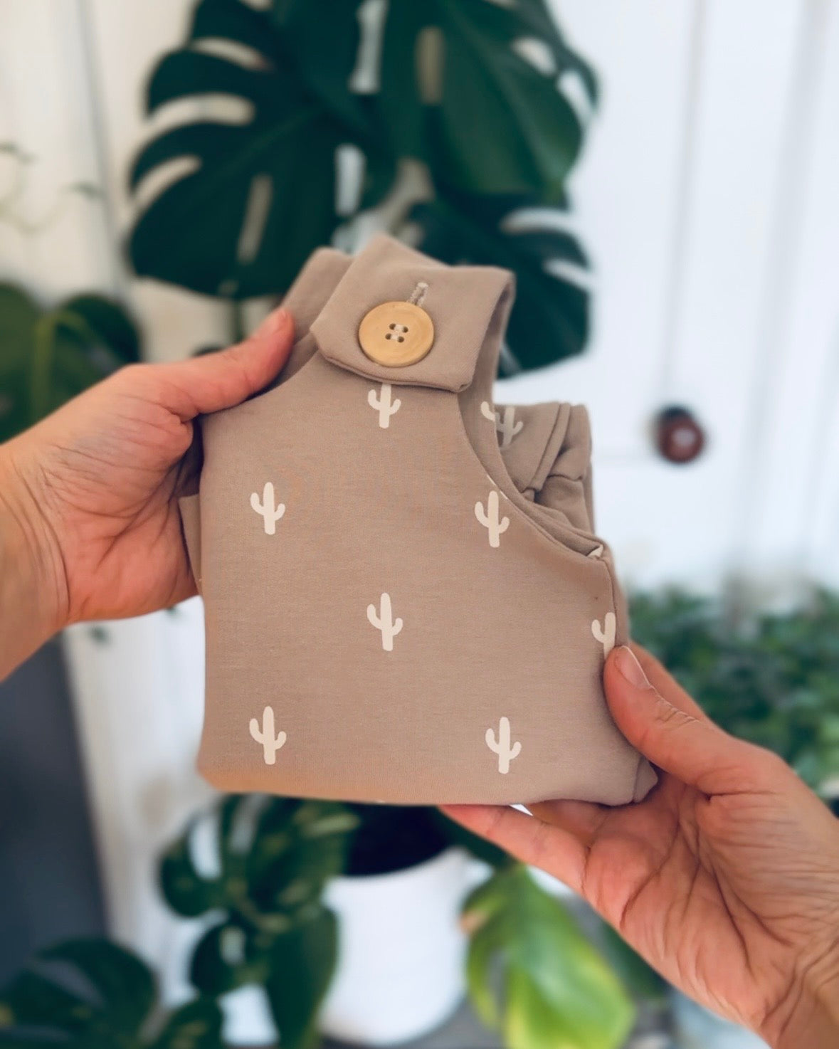 folded gray baby romper with white cactus pattern with wooden buttons held by a hands against a blurred green plant background