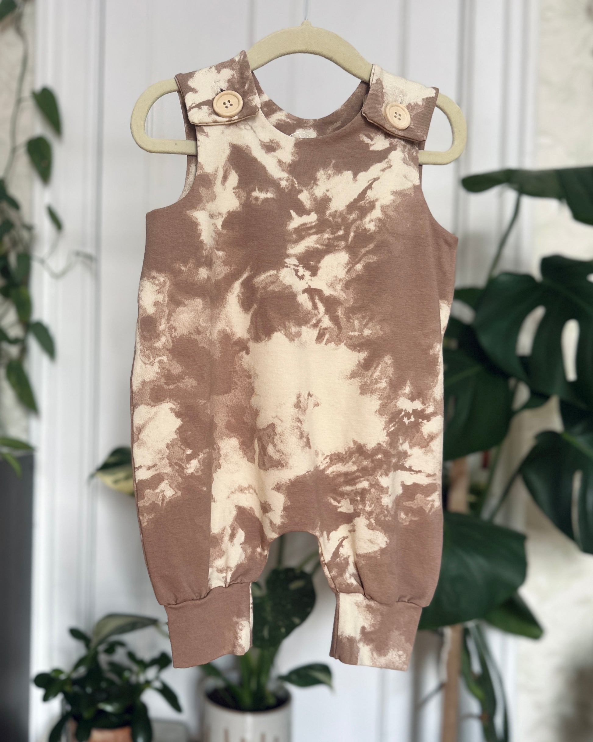 Tie-dye romper with wooden buttons on a hanger with plants in the background