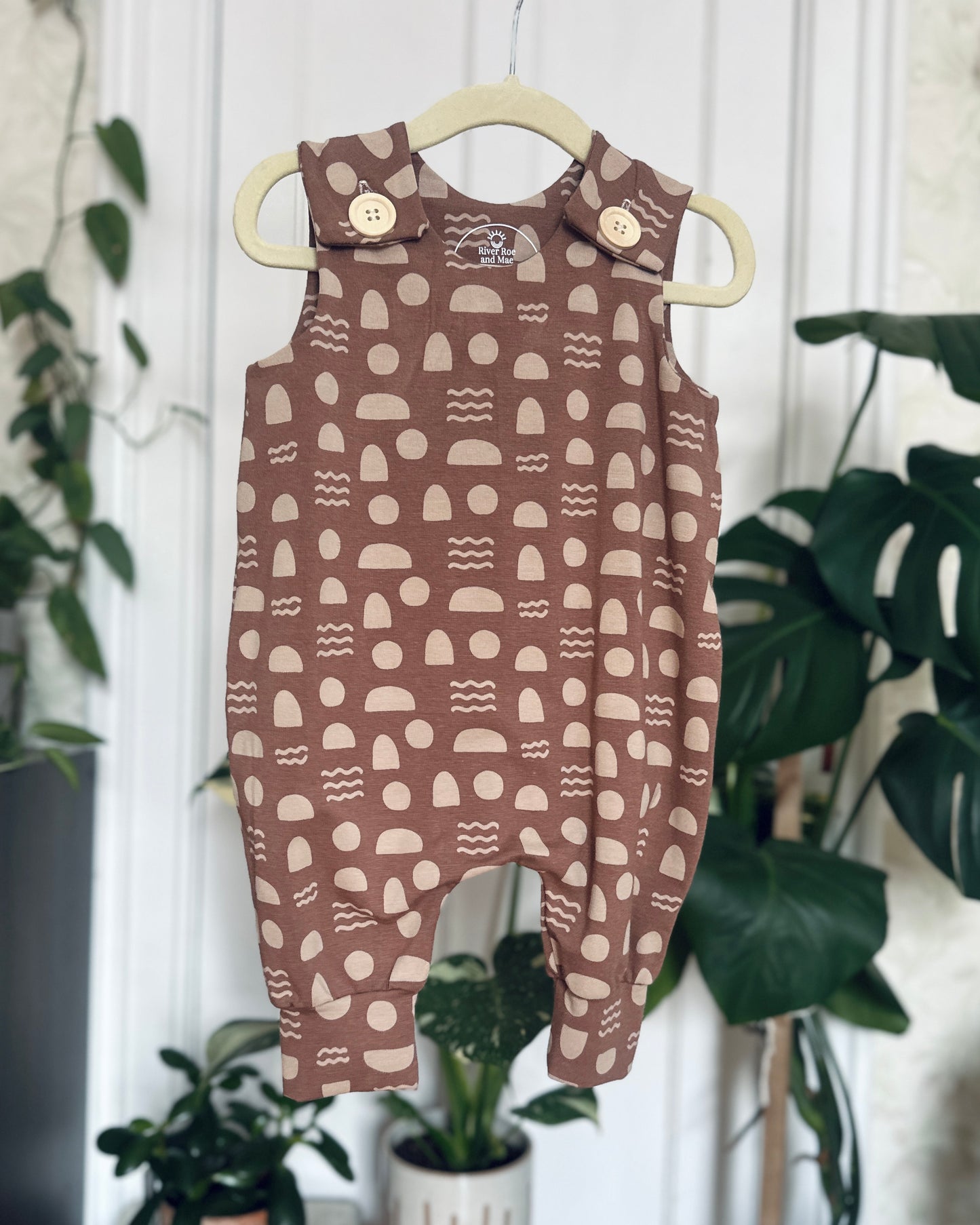 Mauve Geometric Shapes Romper with wooden buttons, hanging on hanger in front of blurred plant background