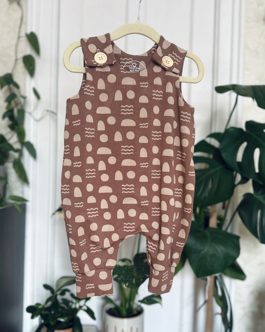 Mauve Geometric Shapes Romper with wooden buttons, hanging on hanger in front of blurred plant background