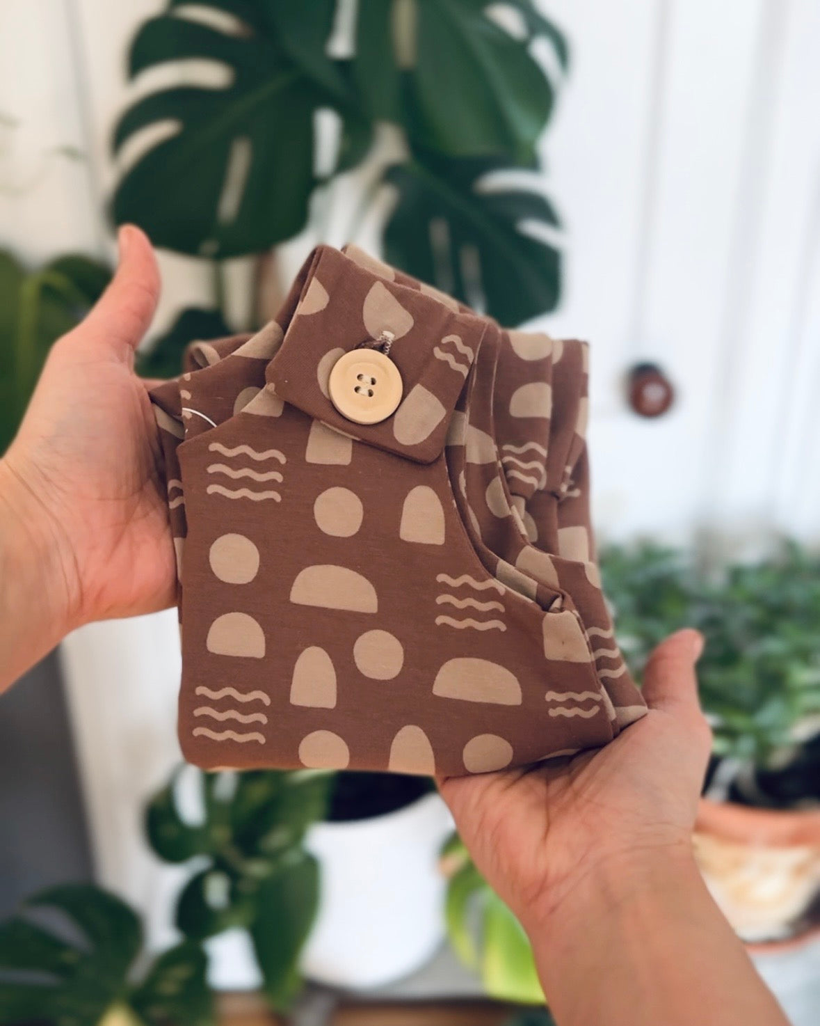 Mocha Shapes room to grow baby romper folded with wooden button at shoulder strap. held by hands in front of a blurred background