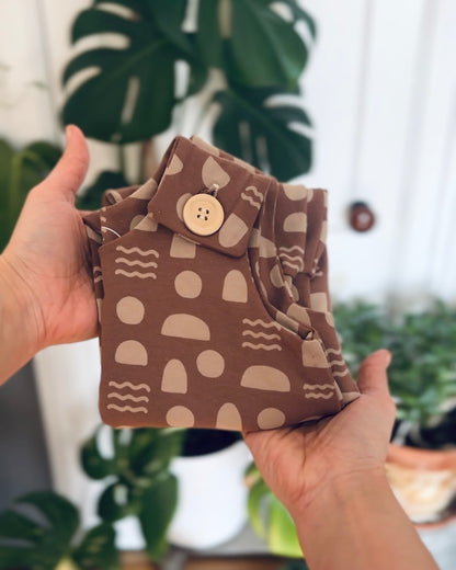 Mocha Shapes room to grow baby romper folded with wooden button at shoulder strap. held by hands in front of a blurred background