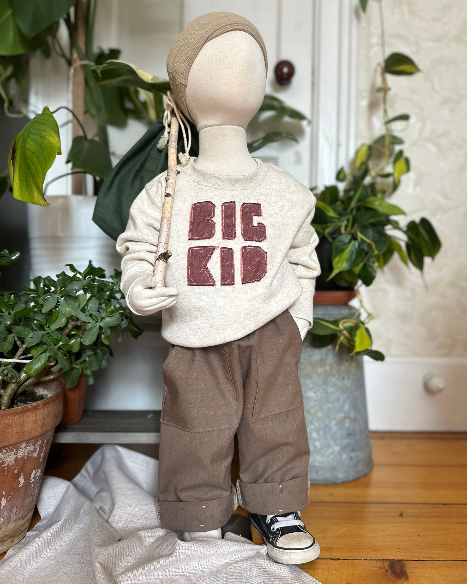 Mannequin wearing a 'BIG KID' sweatshirt and River Rock Caterpillar Pants in a room with plants