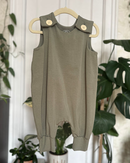 Green baby romper with button details on a hanger against a white wall with plants.